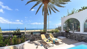Zona de piscina con sillas de descanso, una palmera y vista a la playa.
