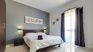 A bedroom with a bed, bedside tables, a wall-mounted light, a window with curtains, and a painting on the wall. - Idyllic Apartment steps from the Promenade (Gzira)