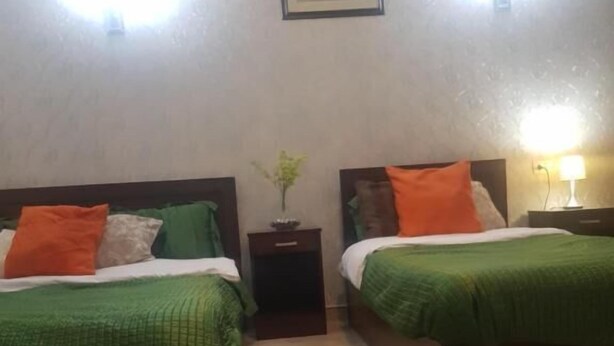 A room with two beds, each with green bedding and orange pillows, a small bedside table with a lamp, and a patterned rug on the floor. - Downtowninn (Cairo)
