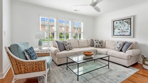 Una sala de estar con un sofá modular beige, una mesa de centro de vidrio, sillas de mimbre y un ventilador de techo.