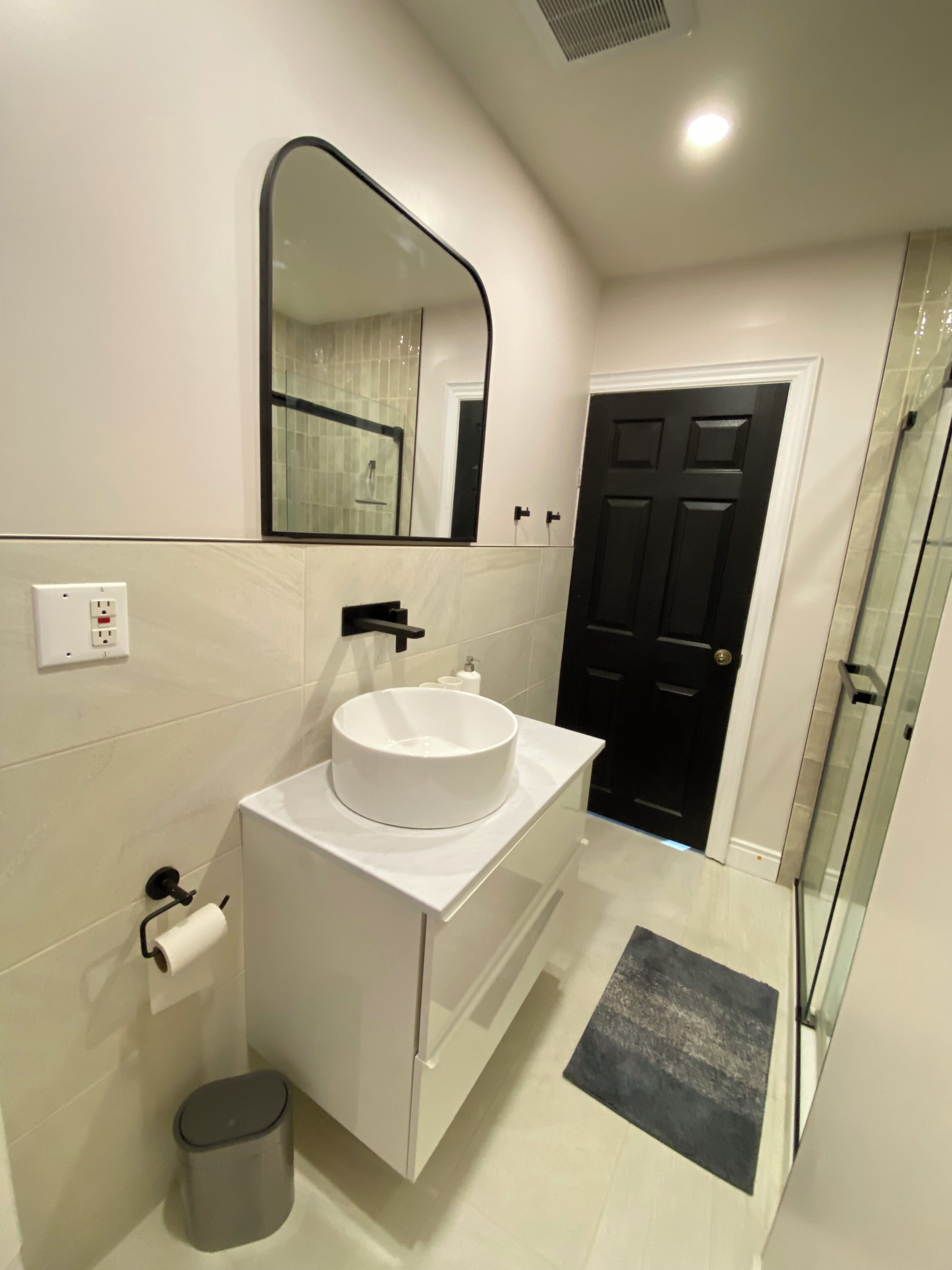A modern bathroom with a white vanity, a large mirror, a black door, and a glass shower enclosure.