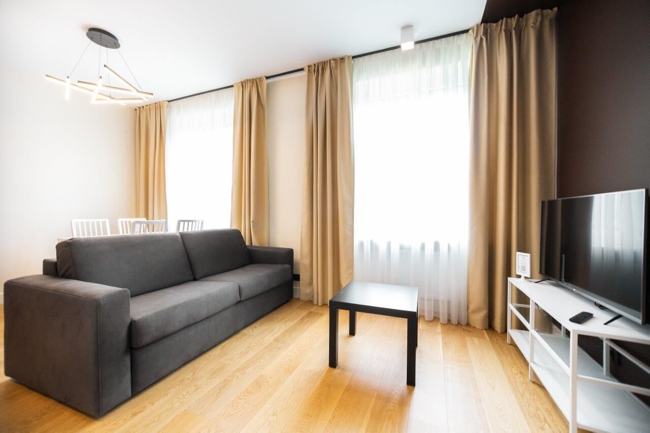 En moderne stue med en grå sofa, et lille bord, et fjernsyn på et hvidt stativ og beige gardiner.