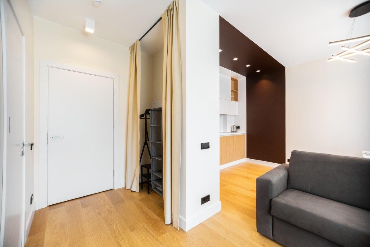 Uma sala de estar moderna com sofá cinzento, soalho de madeira, porta branca e uma área de cozinha ao fundo.