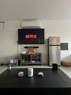 Ein Wohnzimmer mit einem Flachbildfernseher, der „NETFLIX“ in roten Buchstaben anzeigt, einem Kühlschrank mit dem Schild „WELCOME AMIGOS“ und einer kleinen Kommode mit verschiedenen Gegenständen.