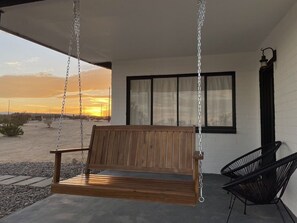 A wooden swing bench under a covered porch with a view of the sunset.