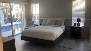 A bedroom with a large bed, a nightstand, a lamp, and a view of a pool through the sliding glass door.