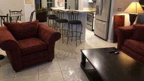 A modern living room with a sofa, a coffee table, and a TV. There is a kitchen area with a refrigerator, microwave, and a dining area with chairs.