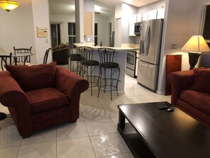 Una sala de estar moderna con un sofá, una mesa de centro y un televisor. Cuenta con una cocina equipada con refrigerador, microondas y un comedor con sillas.