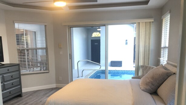 A bedroom with a large bed, a dresser, a window with blinds, and a view of a pool through a sliding glass door.