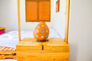 Une malle en bois fermée à clé, posée sur un lit recouvert de draps blancs, près d’une fenêtre ornée de volets en bois.