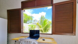 Um espaço de trabalho com um laptop sobre uma mesa de madeira, uma cadeira de plástico branca e uma janela com persianas castanhas.