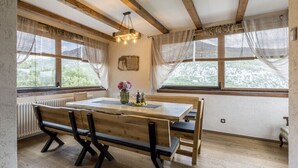 Een eethoek met een houten tafel en banken, grote ramen met gordijnen, een hanglamp en uitzicht op groen buiten.