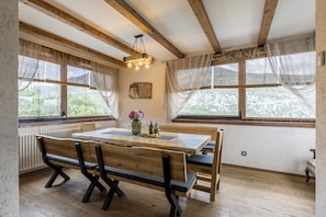 A dining area with a wooden table and benches, large windows with curtains, a hanging light fixture, and a view of greenery outside. - vila posjeduje jacuzzi, saunu s tušem, grijani bazen ..... (Omiš)