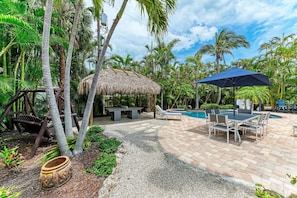 A tropical outdoor area with a pool, lounge chairs, a thatched-roof structure, and a dining table.