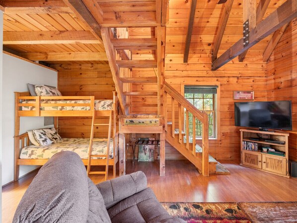 Interior de una cabaña de madera con literas, una escalera, un televisor y una estantería.