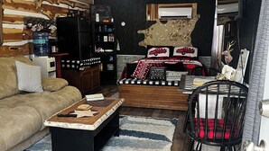 Ein gemütliches Wohnzimmer mit einer Couch, einem Couchtisch und einem gemusterten Teppich. Es gibt eine Küchenzeile mit einem Kühlschrank und einem Wasserspender. An der Wand ist eine Dekoration mit Hirschkopf zu sehen.