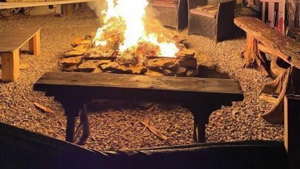 Ein Lagerfeuer mit einem Holztisch in der Mitte, umgeben von Sitzgelegenheiten und einem Gebäude mit der Aufschrift "The Wood Shed".