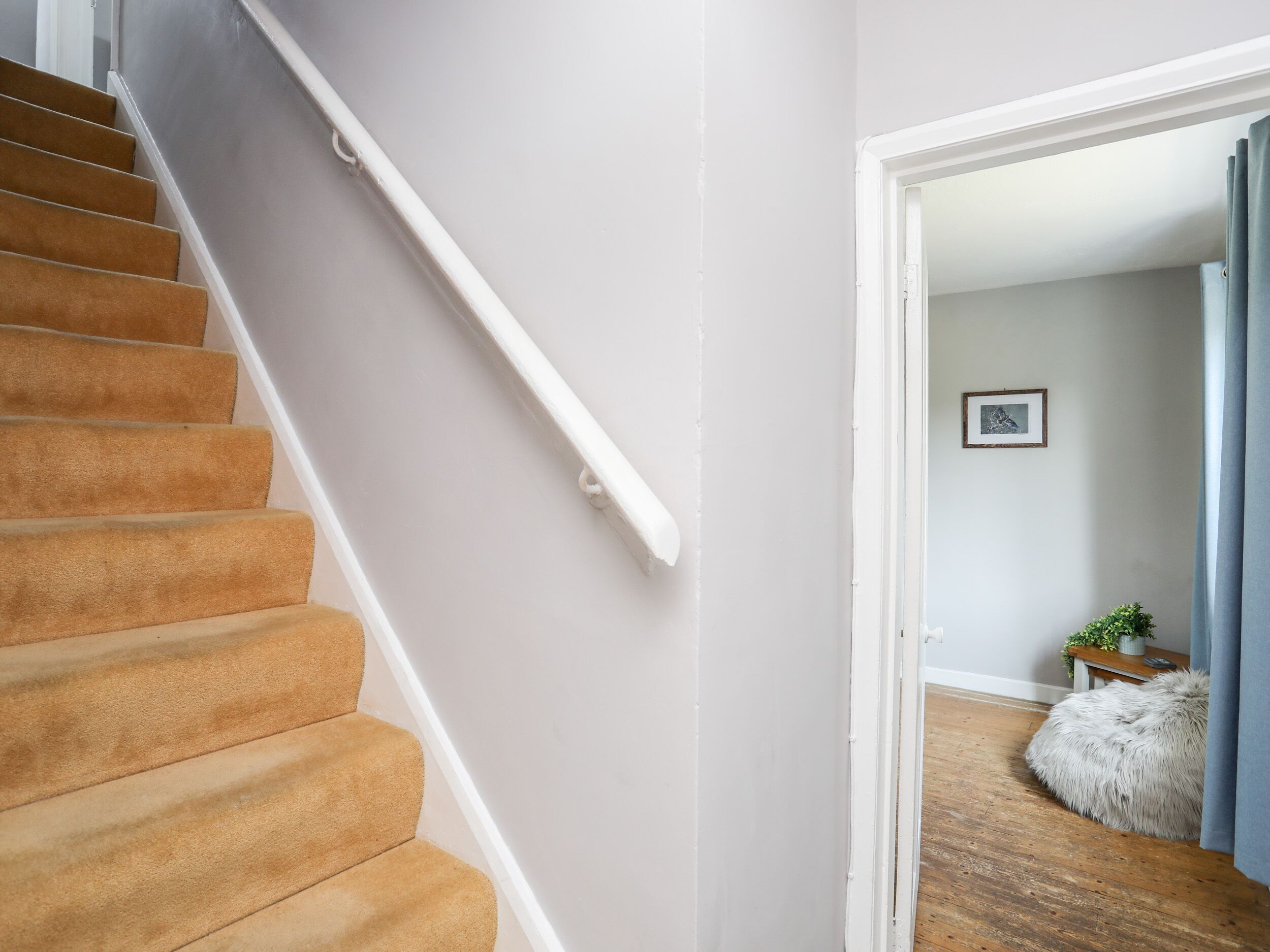 Un escalier dont les marches sont recouvertes d’une moquette beige, menant à une pièce dotée d’un plancher en bois, d’un petit tapis rond blanc et d’un tableau encadré accroché au mur.