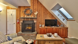 A cozy living room with a fireplace, wooden walls, a television, and a skylight. - The Sky's the Limit at Mountain Green 2BD/2BA: Close to Slopes / Discount Tickets (Killington)