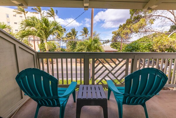 A balcony with two green chairs and a small table, overlooking a street with palm trees and buildings. - Great rates at Kihei Bay Surf G259 Comfortable Studio Space! (Kihei)