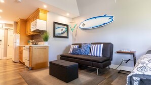 A compact living space with a kitchenette, a sofa with patterned cushions, a surfboard decoration, and a small side table. - Great rates at Kihei Bay Surf G259 Comfortable Studio Space! (Kihei)