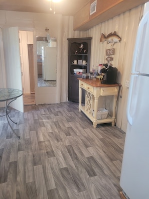 A kitchen with a wooden floor, a white refrigerator, a wooden table with a basket, and a dark cabinet with shelves.