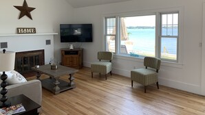 Un salon avec une cheminée, un téléviseur, deux fauteuils à motifs et une grande fenêtre donnant sur une étendue d’eau.