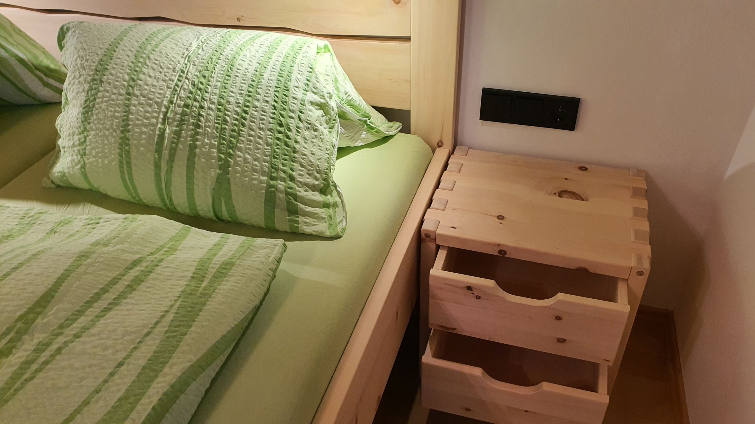Una cama con ropa de cama a rayas verdes, una mesita de noche de madera con cajones y un enchufe en la pared.