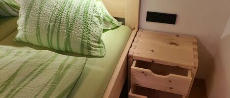 Un lit avec une literie à rayures vertes, une table de chevet en bois avec des tiroirs et une prise électrique fixée au mur.