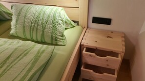 Uma cama com roupa de cama verde listrada, uma mesinha de cabeceira de madeira com gavetas e uma tomada na parede.