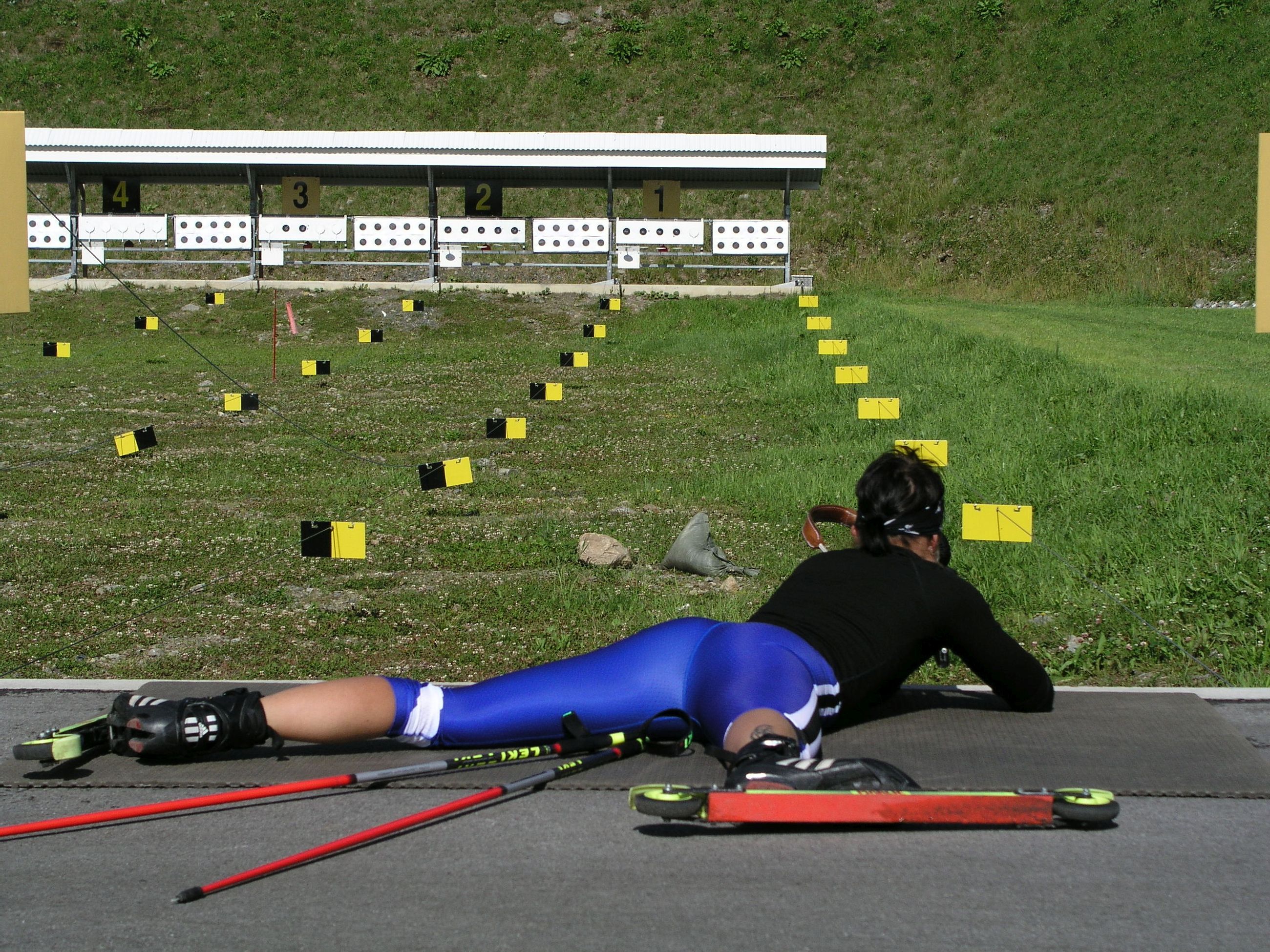 A person lying on the ground with skis and poles, a numbered barrier, and a grassy hill in the background.