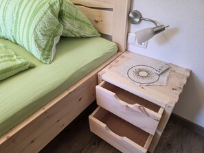 En træseng med grønt sengetøj og puder, en sengetlampe og et natbord i træ med skuffer.