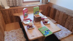 En träbänk med blommiga kuddar, ett bord med en bok, en kopp, en sked och en liten trälåda.
