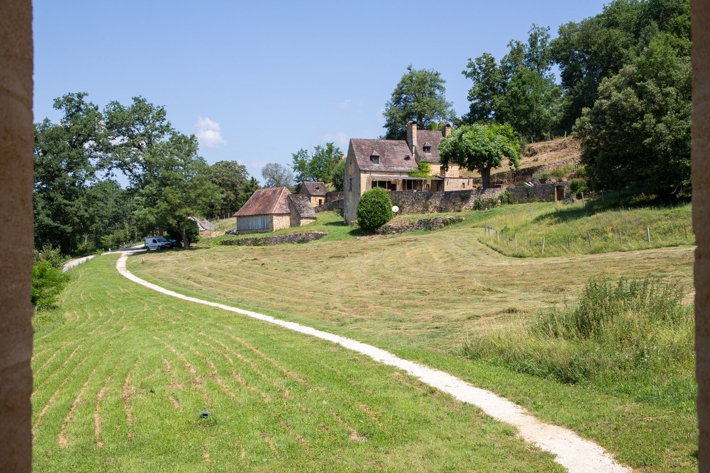 Un paysage rural avec un chemin de terre menant à une maison en pierre dotée d’un toit en pente, entourée d’arbres et de champs ouverts.