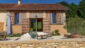 A stone building with a tiled roof, a wooden door, and a small porch with two chairs and a table. - La Grange Milou (Beynac-et-Cazenac)