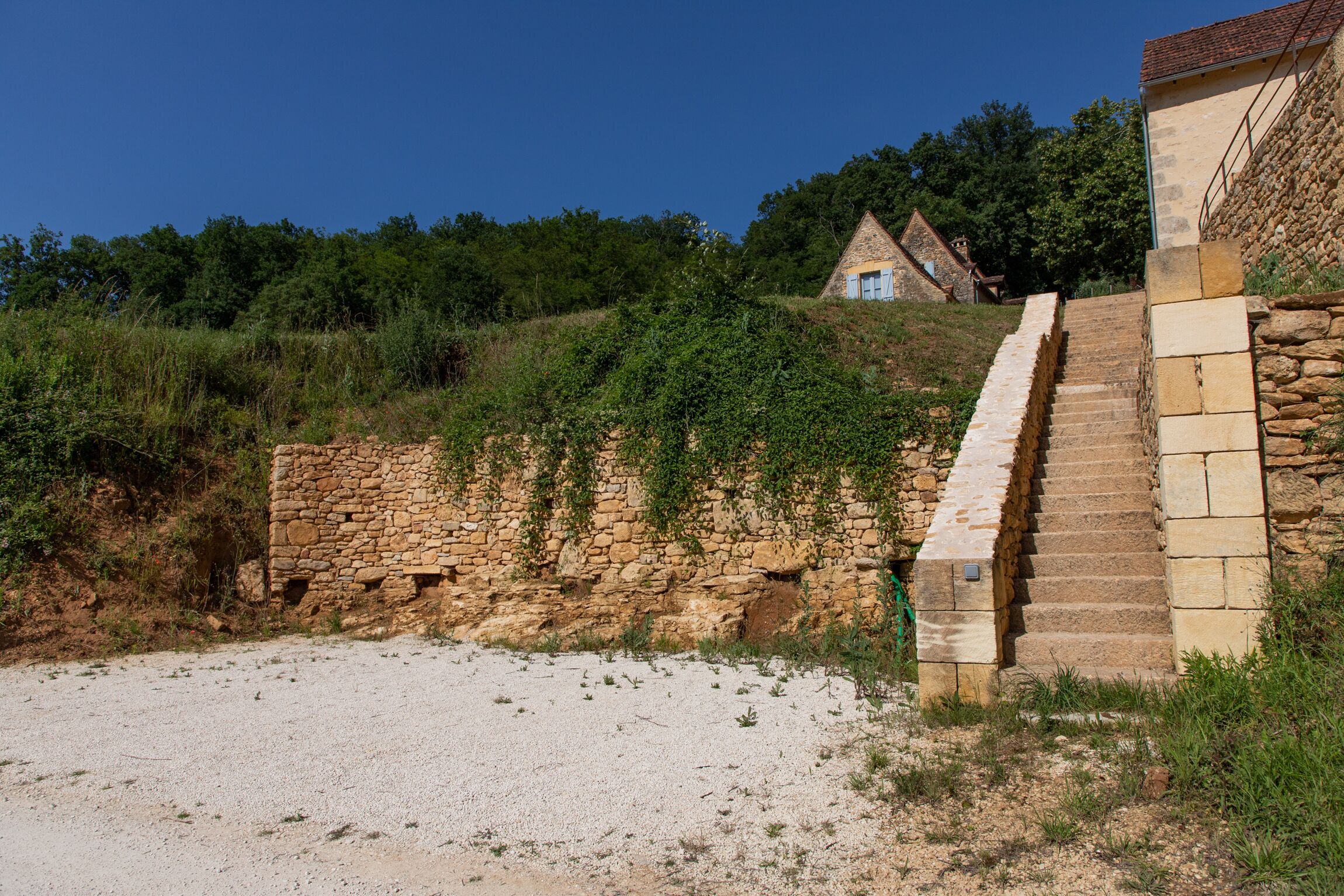 A stone staircase leading to a traditional stone building with a sloped roof, surrounded by greenery and a clear sky.