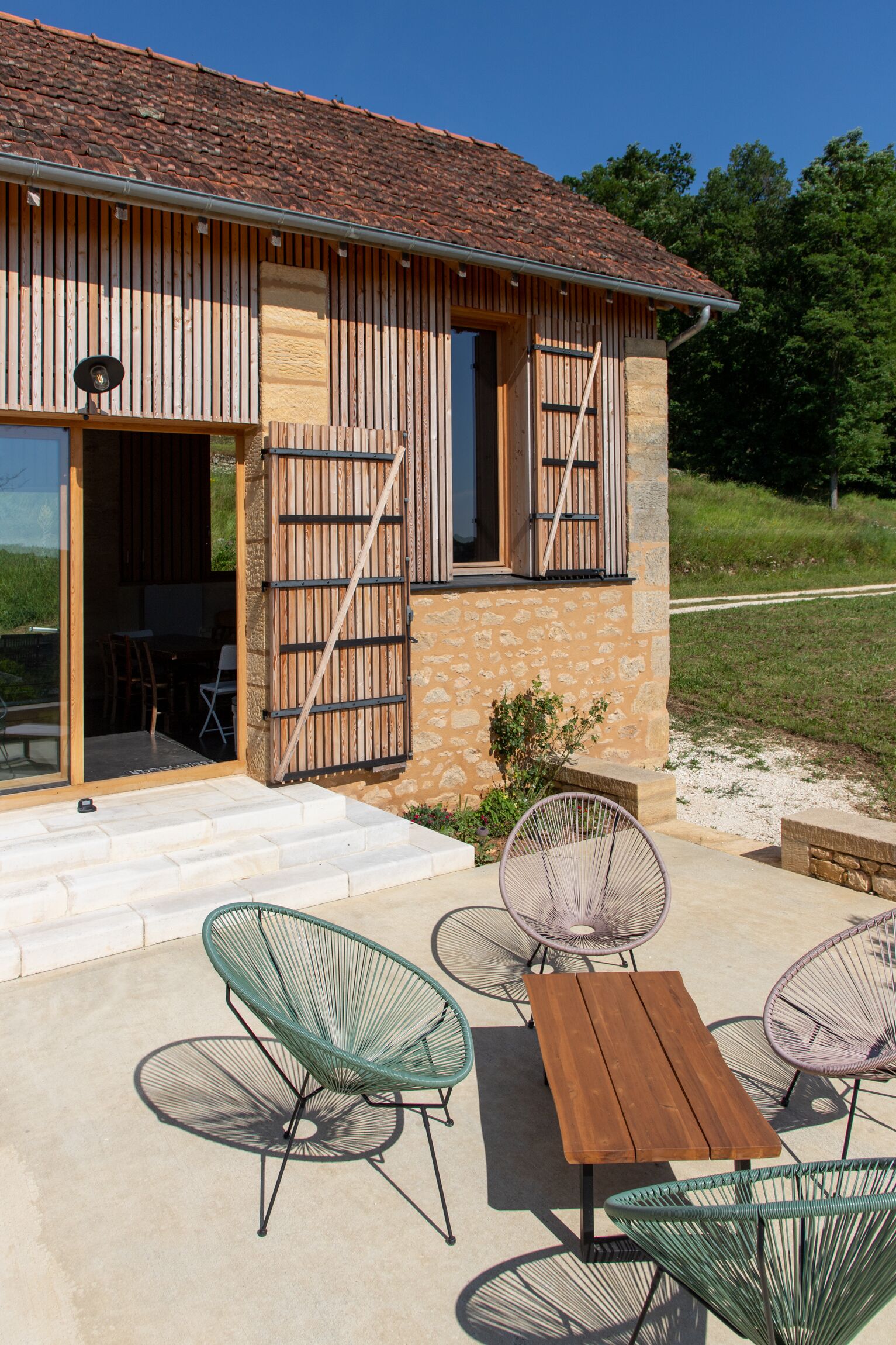 A rustic building with a stone facade, wooden shutters, and a patio with metal chairs and a wooden table.