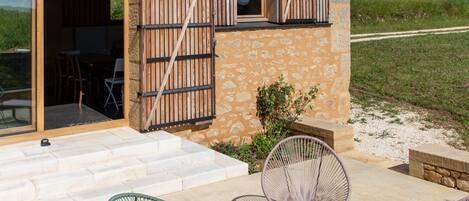 A rustic building with a stone facade, wooden shutters, and a patio with metal chairs and a wooden table.