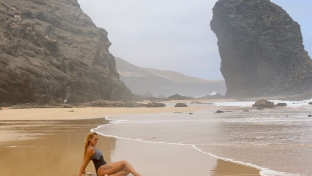 A person sitting on a sandy beach with large rock formations in the background. - Olas de Lobos (Corralejo)