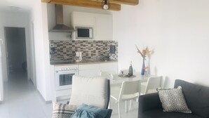 A modern kitchen with white cabinets, a microwave, and a tiled backsplash. There is a dining area with white chairs and a table, and a living room with a dark sofa and patterned cushions. - Olas de Lobos (Corralejo)