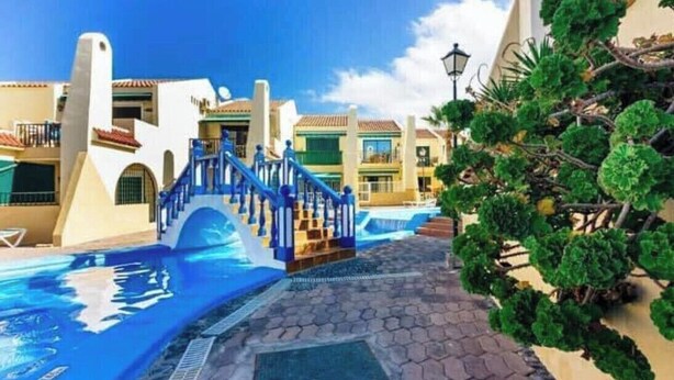 A swimming pool with a blue staircase leading to a covered area, surrounded by buildings with tiled roofs and a clear sky. - A&V Mareverde Costa Adeje Luxury Poolfront Apartment (Costa Adeje)