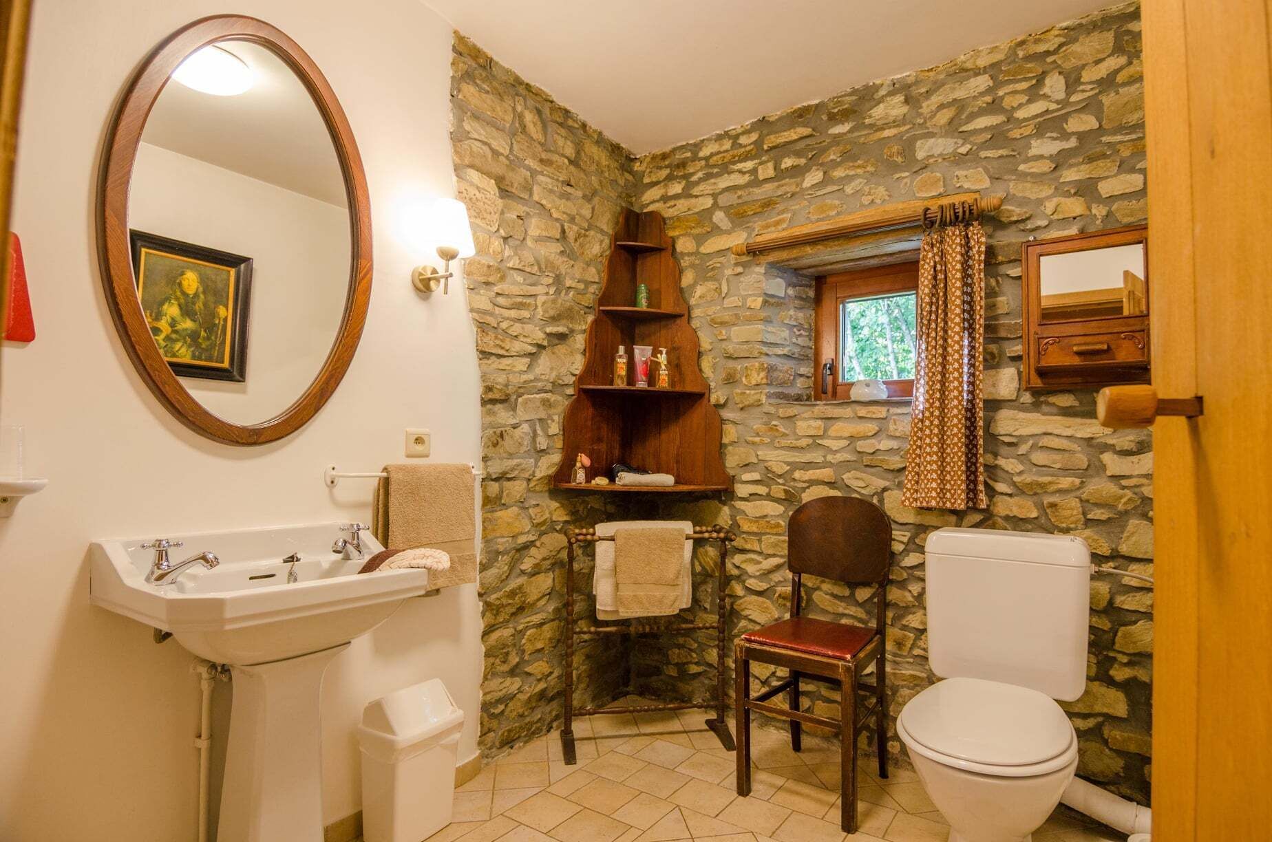 Une salle de bain avec un mur en pierre, une chaise en bois, des toilettes blanches et un lavabo avec un miroir.