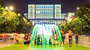A large building with classical architecture, a fountain in the foreground, and people sitting on benches. - Birds of Paradise (Bucharest)