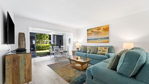 A modern living room with a sofa, coffee table, and TV. There is a dining area with chairs and a table visible through an open door. - Siesta Dunes 6-102, Peaceful Garden View Unit, 2br/2ba, 1st Floor Condo (Sarasota)
