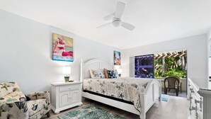 A bedroom with a bed, nightstands, a chair, a ceiling fan, and a window with a view of greenery. - Siesta Dunes 6-102, Peaceful Garden View Unit, 2br/2ba, 1st Floor Condo (Sarasota)