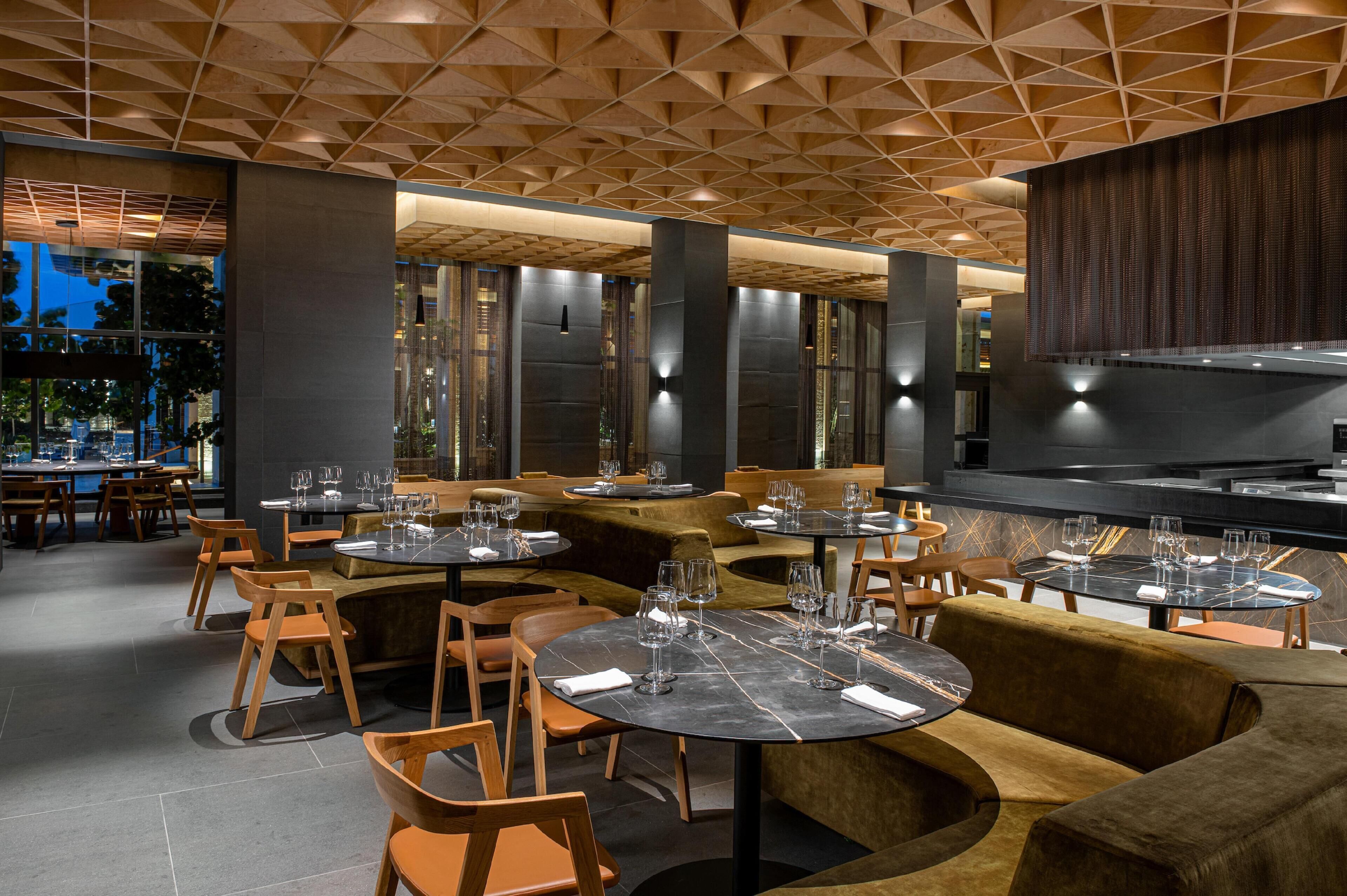 A modern restaurant interior with round tables, cushioned seating, and a geometric ceiling design.