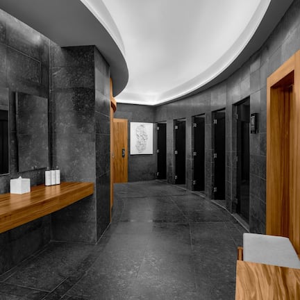 A modern bathroom with dark tiled walls, wooden benches, and a long counter with sinks.
