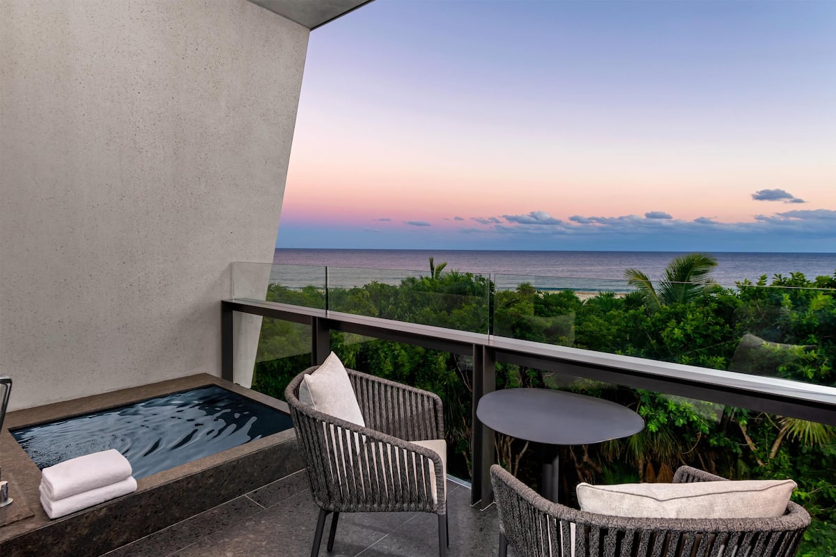 Un balcon doté d’un jacuzzi, de fauteuils en osier et d’une table offrant une vue sur la plage au coucher du soleil.