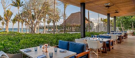 Un área de comedor al aire libre con mesas y sillas, con vista a una playa con palmeras y un edificio de techo de paja.