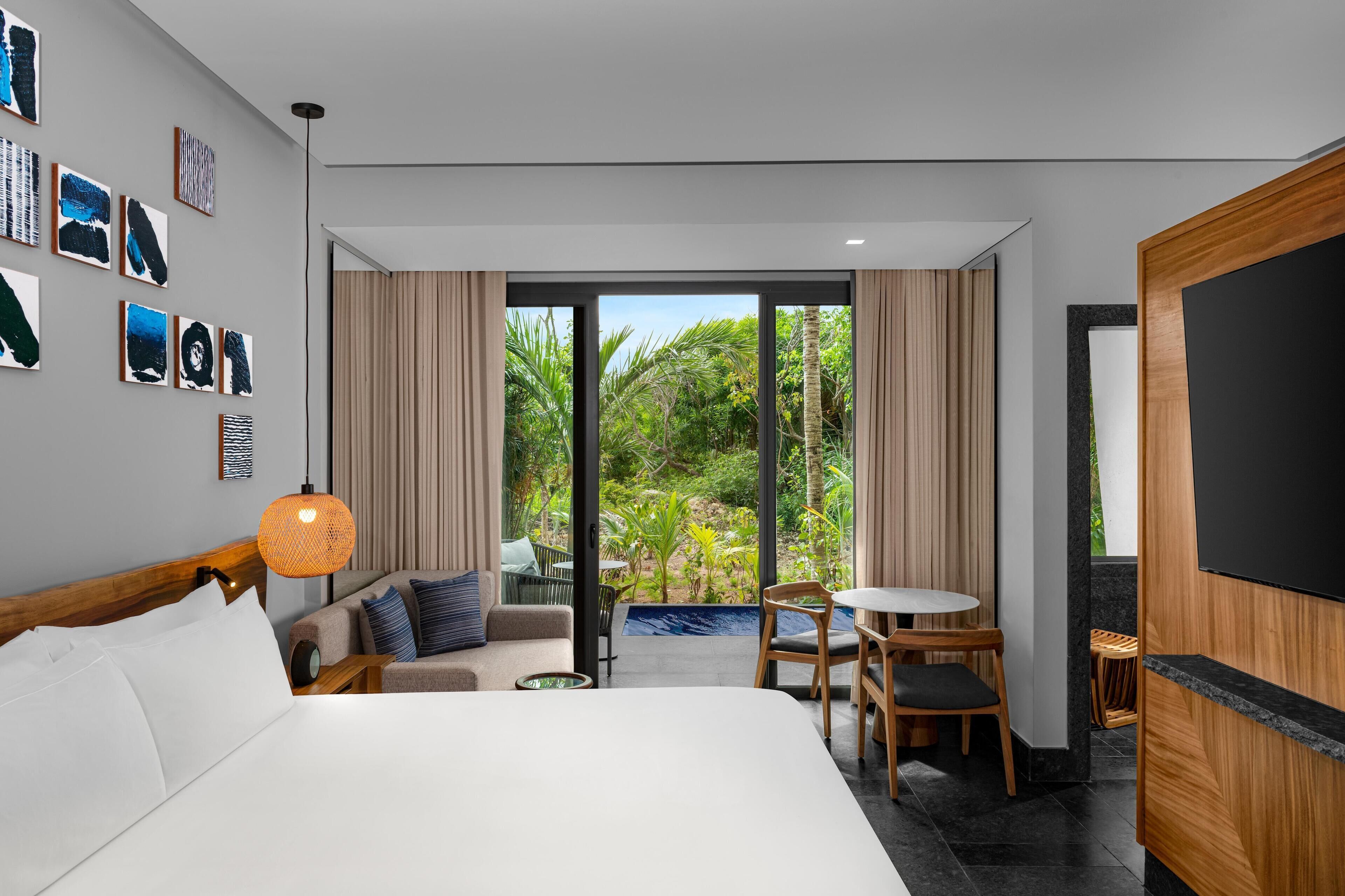 Een moderne hotelkamer met een groot bed, een kleine zithoek en een balkon met uitzicht op groen.